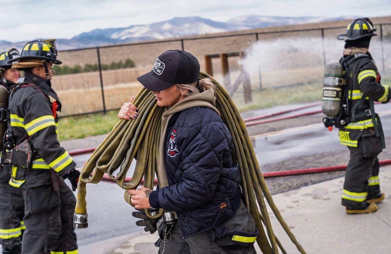 A woman carries a coiled hose load over her shoulder across a wet training ground while a crew works behind her
