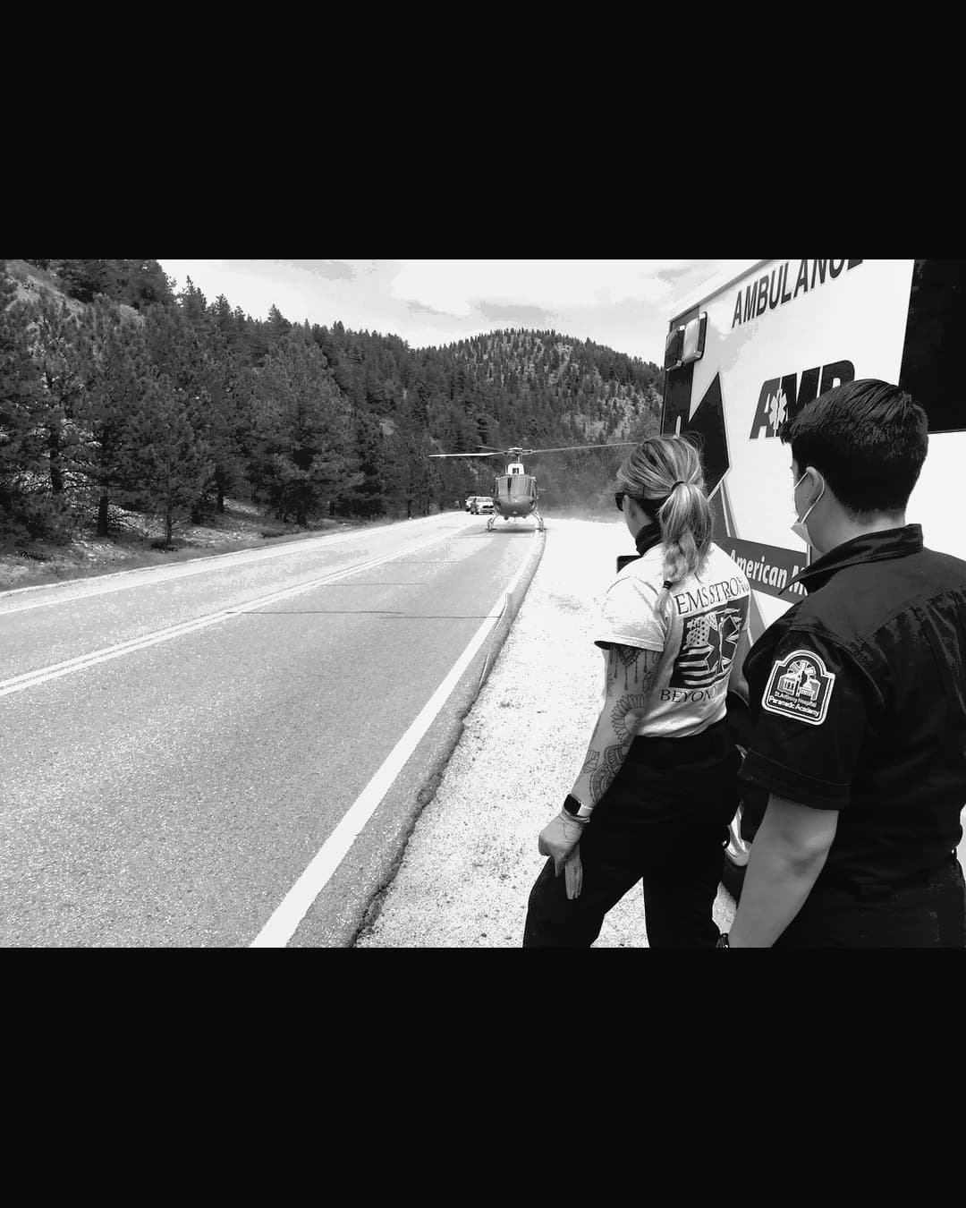 Two EMS providers stand beside an ambulance watching a medical helicopter land on a mountain highway